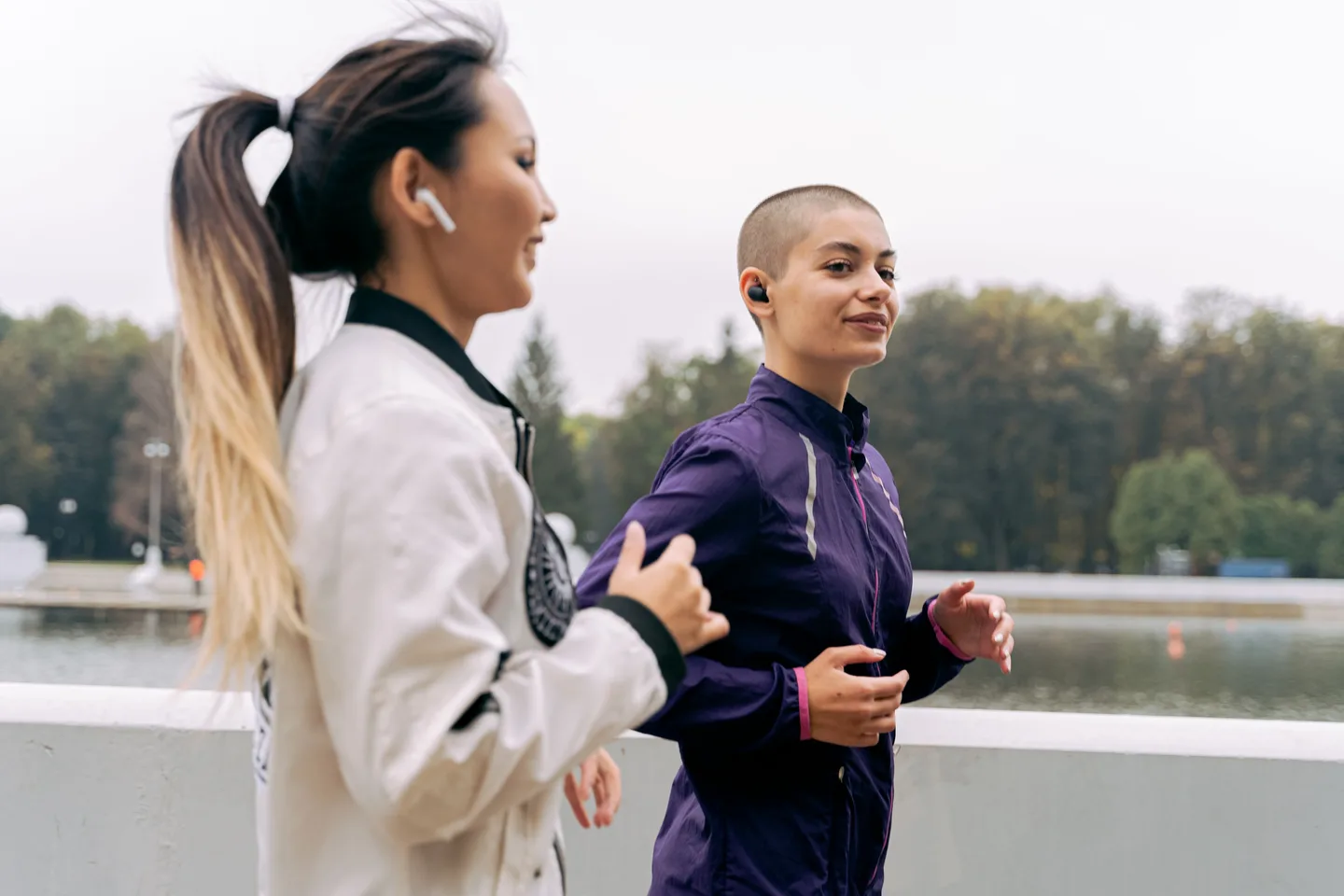 Two friends run side by side beside the water, each wearing earbuds.